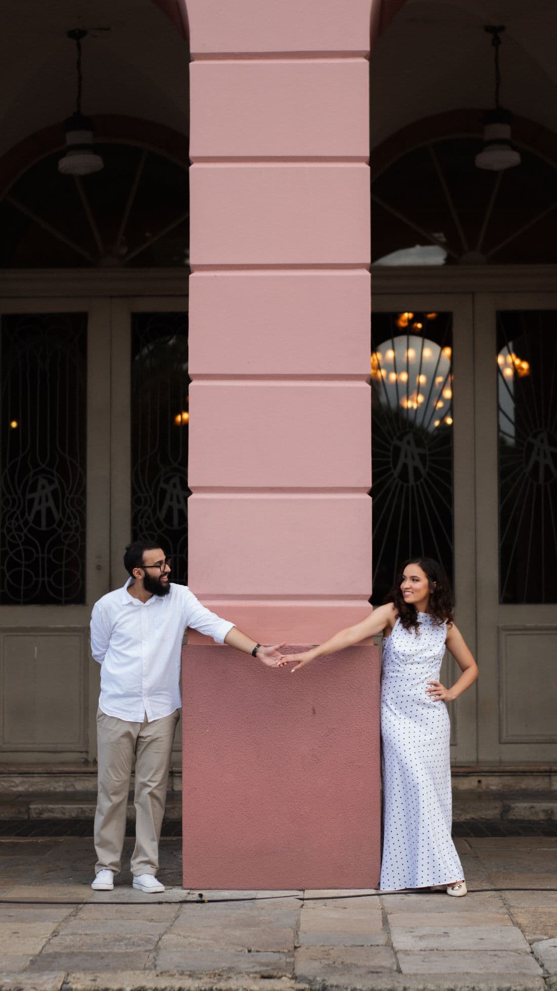 Gabriel e Geovanna, um de cada lado de uma coluna rosa, de mãos dadas